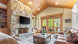 A cozy living room with a stone fireplace, wooden ceiling, and a large window offering a view of greenery. - Town Pointe by KBM (Park City)