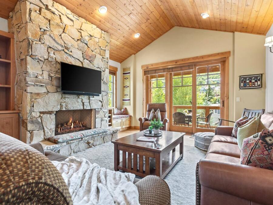 Un salon chaleureux avec une cheminée en pierre, un plafond en bois et une grande fenêtre donnant sur un paysage verdoyant.