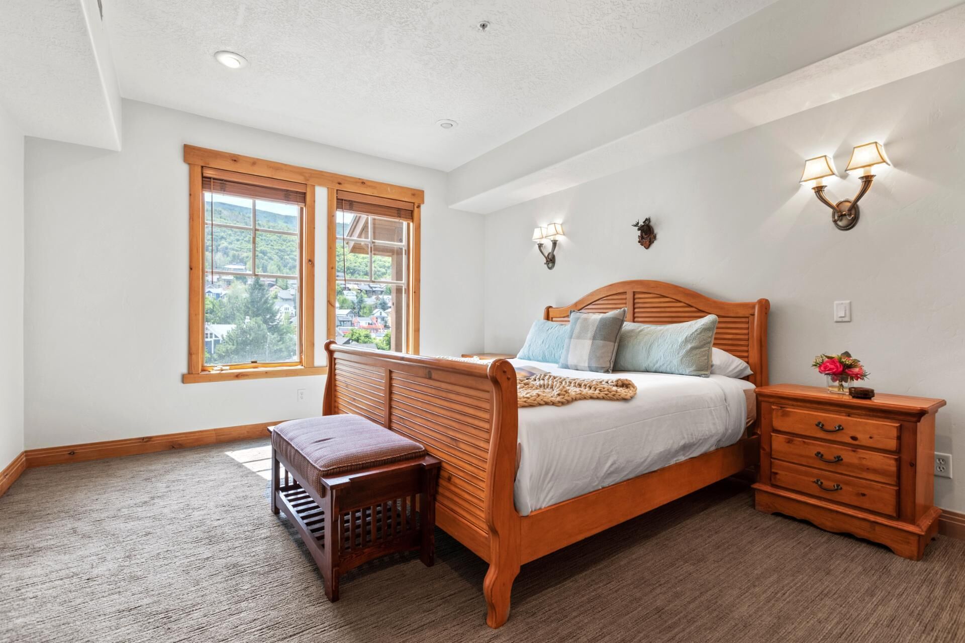 A bedroom with a wooden bed, two bedside lamps, a nightstand, a bench, and a window with a view of houses.