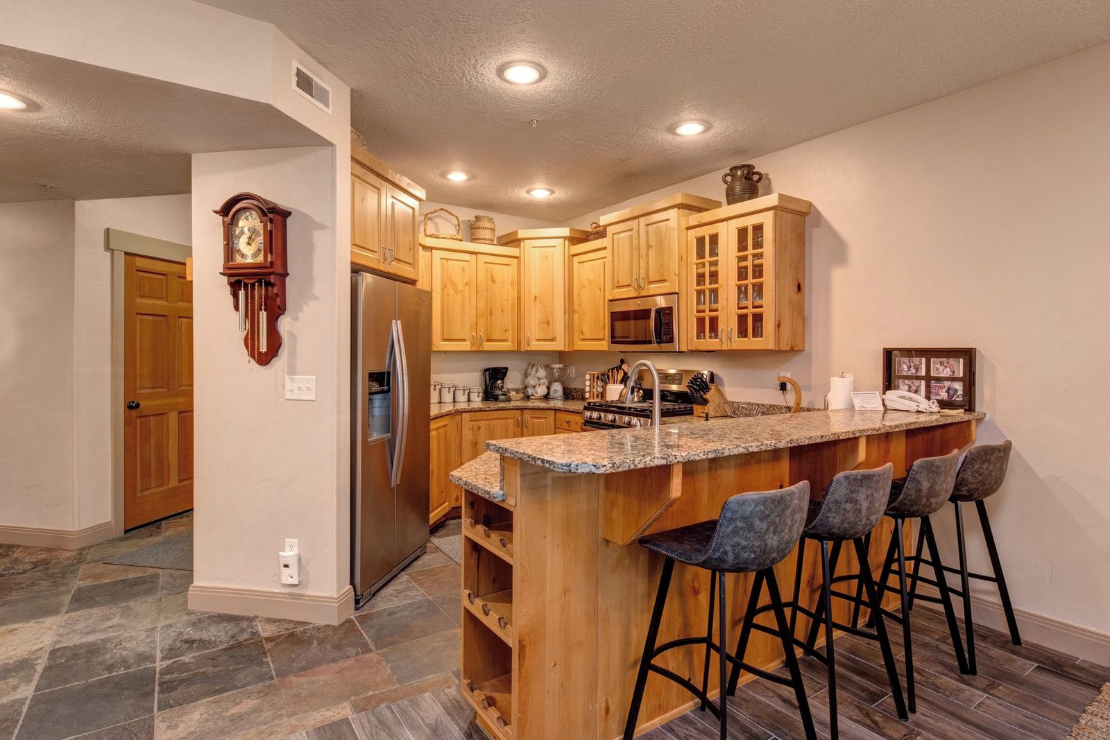 A kitchen with wooden cabinets, a stainless steel refrigerator, a granite countertop, and a bar area with stools.