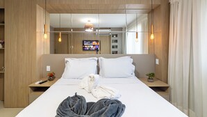 A modern hotel room with a large bed, wooden paneling, a TV, and a bedside table with a remote control. - Cond. Barra Bali | Frente ao Mar | Destino BSM 108 (Barra de São Miguel)