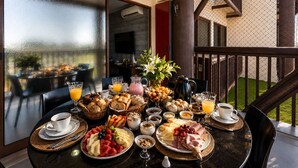 A breakfast buffet with various foods and beverages on a table, set on a balcony with a view of a building and a garden. - Cond. Barra Bali | Frente ao Mar | Destino BSM 108 (Barra de São Miguel)