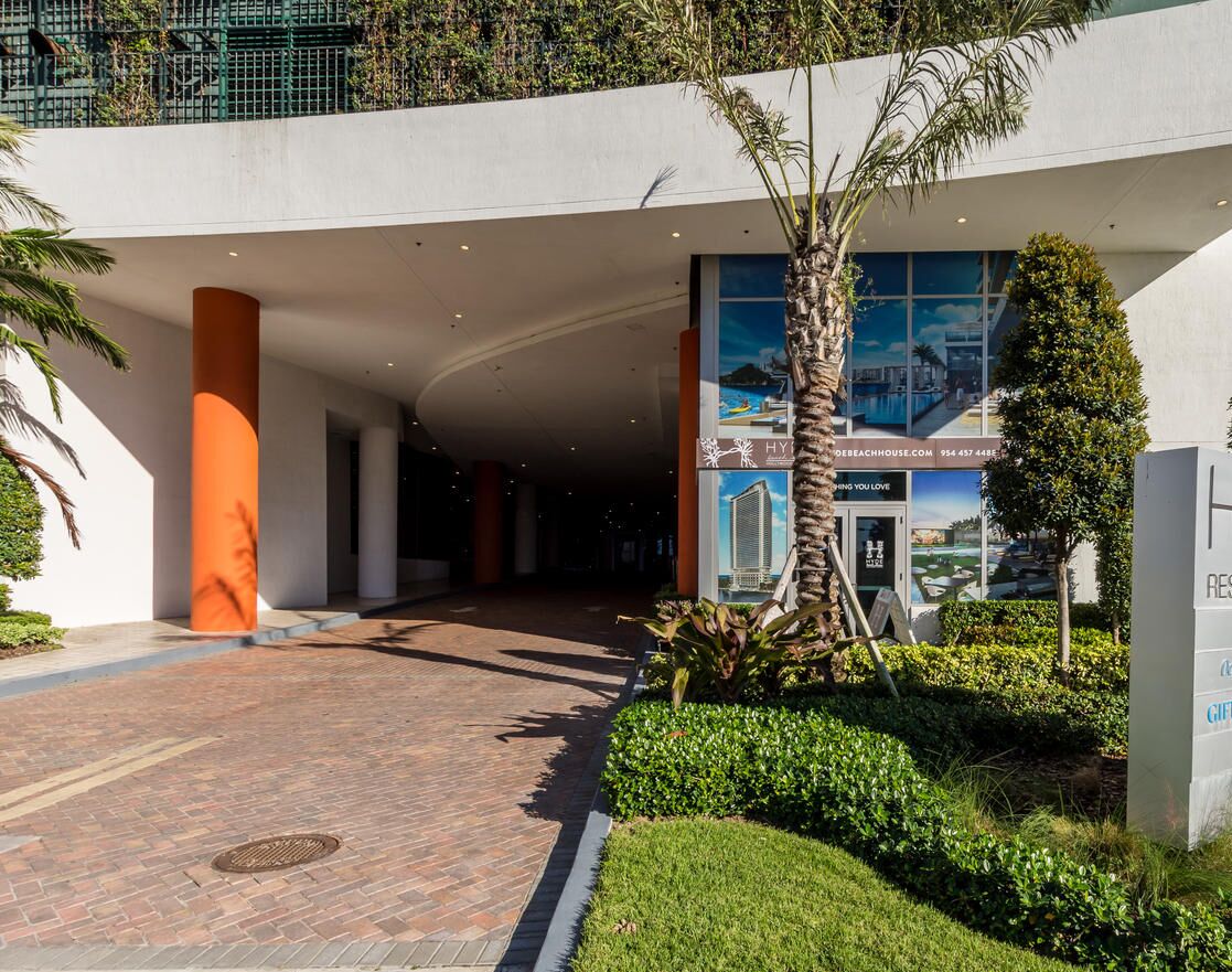 A modern building entrance with a curved pathway, palm trees, and a large glass window displaying a beach scene.