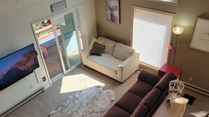 Ein Wohnzimmer mit einem beigefarbenen Sofa, einem braunen Sofa, einem weißen, flauschigen Teppich, einem Holztisch und einem Fernseher links.
