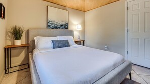 Un dormitorio con cama, mesita de noche, lámpara, planta en maceta y una puerta.