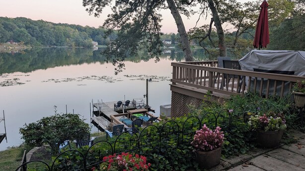 A lakeside deck with outdoor seating and a covered area, surrounded by lush greenery and a calm lake. - Tranqulity and relaxtion destination (Syracuse)