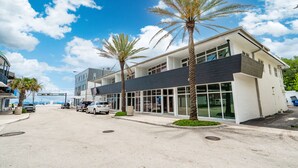 A modern building with large windows and a flat roof, surrounded by palm trees and parked cars. - Purple Pelican by Opus (Suite 4) (Fort Lauderdale)