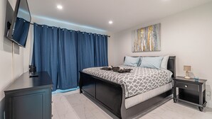 A bedroom with a bed, a nightstand, a TV, and a painting on the wall. - Purple Pelican by Opus (Suite 4) (Fort Lauderdale)
