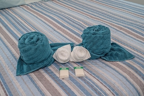 A bed with a striped blue and beige bedspread, two rolled-up towels, and two bottles of face and body bar soap.