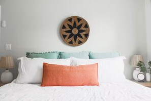 A bed with white bedding and a single orange pillow, flanked by two lamps and a wall-mounted decorative item.