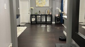 Um corredor com assoalho de madeira escura, uma porta branca, um console preto com objetos decorativos e um espelho grande refletindo o ambiente.