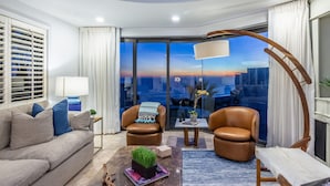 Una sala de estar moderna con un sofá, dos sillones de cuero, una mesa de centro con una planta y vistas de la ciudad al atardecer.