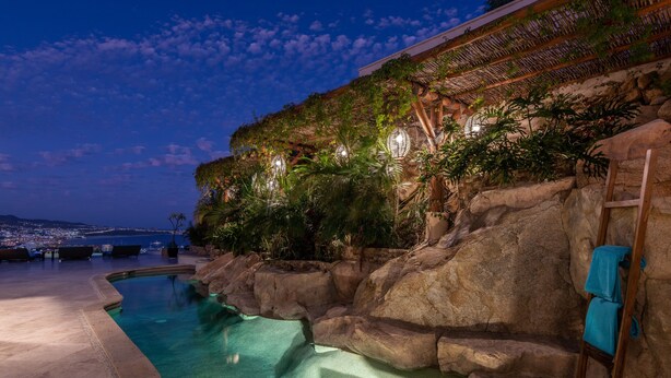 Una piscina de aguas cristalinas, rodeada de rocas y palmeras, con vista a la ciudad al fondo.