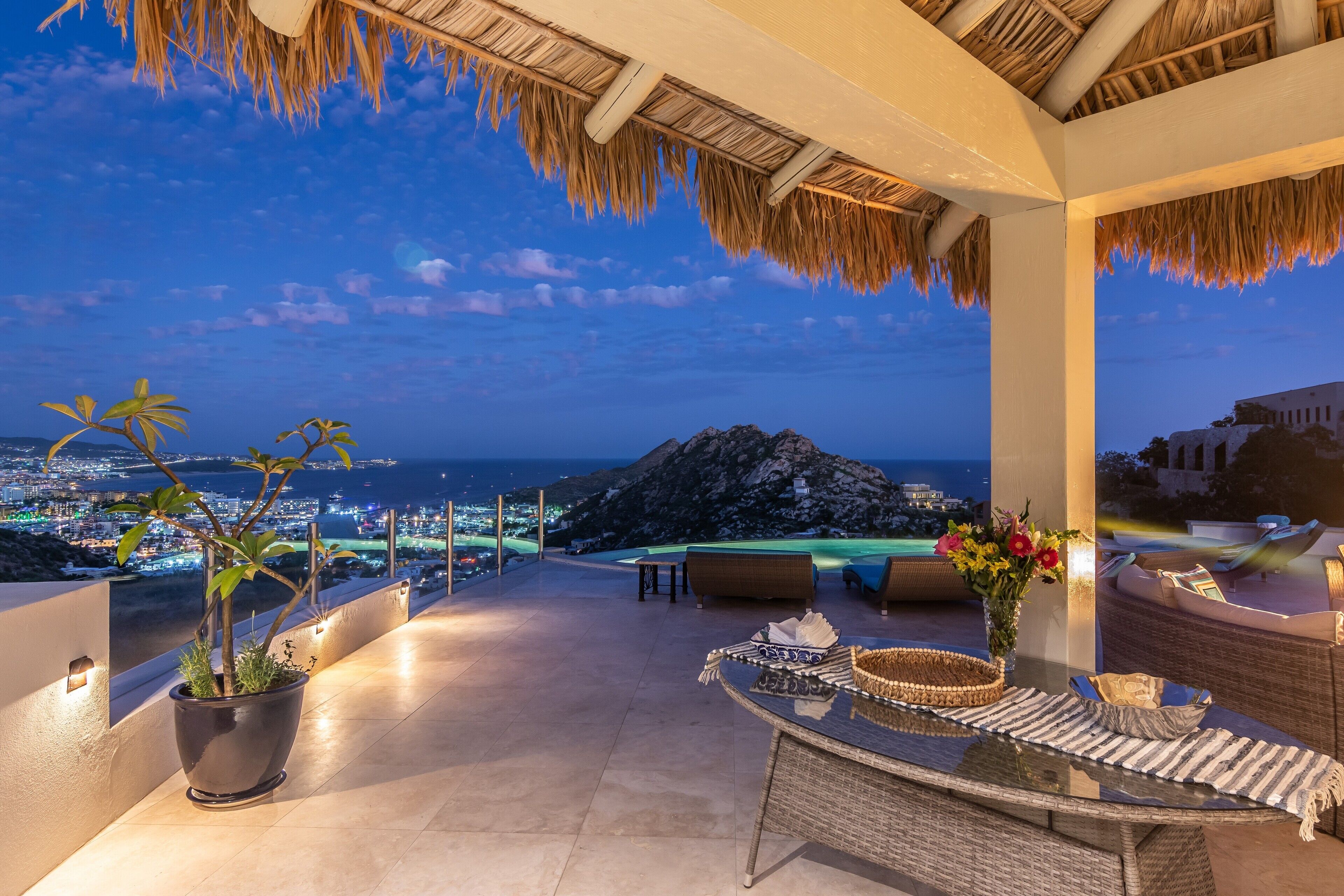 A rooftop terrace with a view of the city and mountains, furnished with a table and chairs, and potted plants.