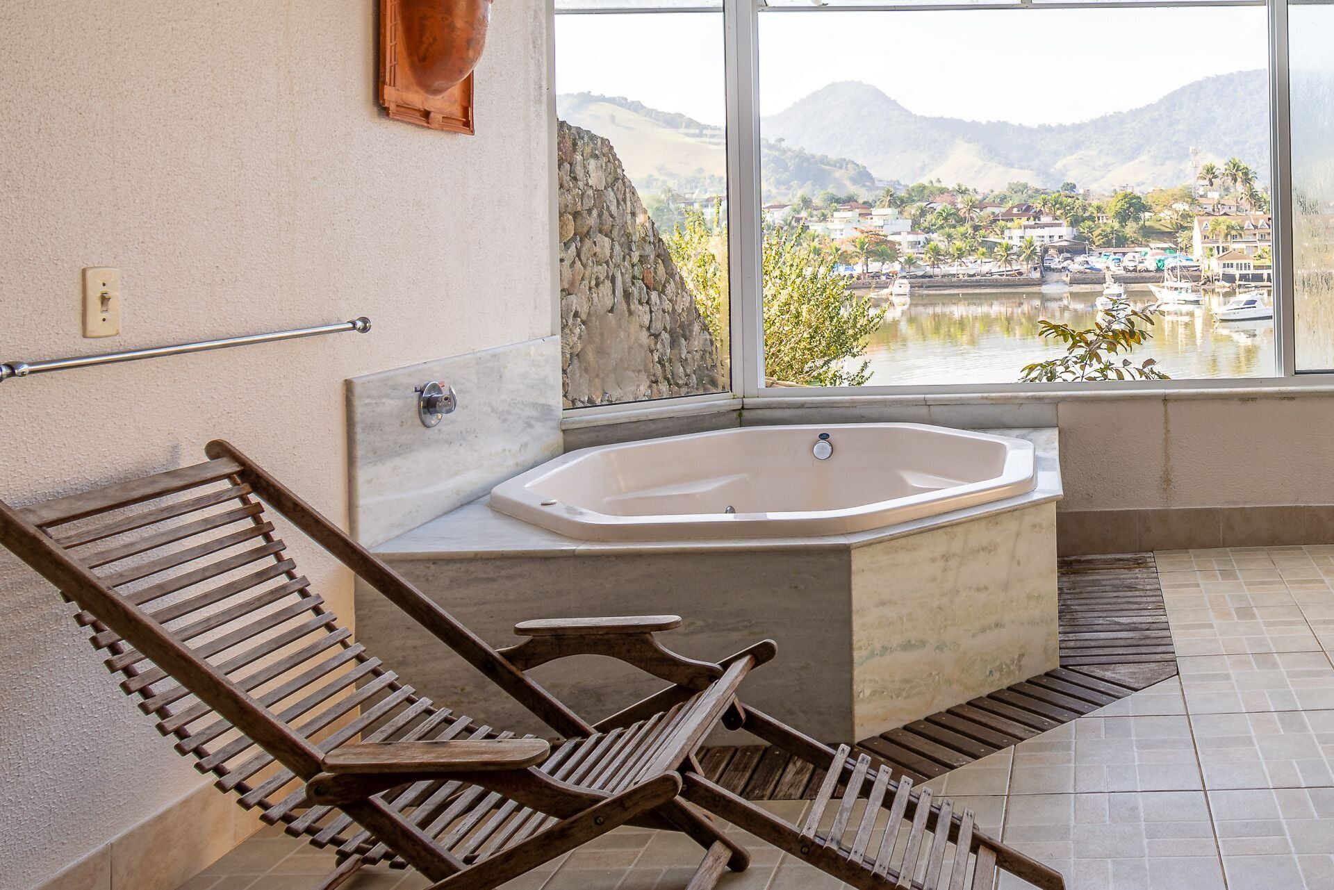 A bathroom with a bathtub, wooden chairs, a stone wall, and a view of a lake and mountains.