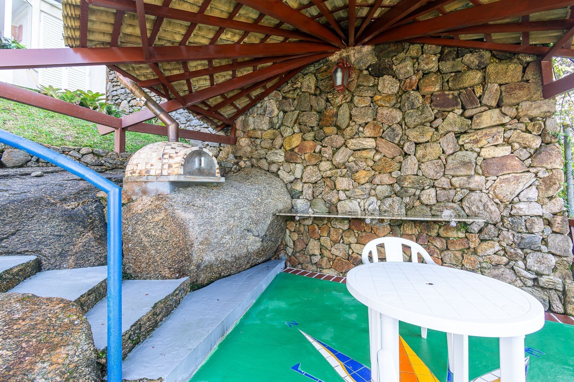 A stone wall with a stone oven, a white table, and a blue chair under a wooden roof structure.