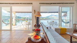 Een ruime woonkamer met uitzicht op een meer en bergen, met een hangmat, een bank en een eettafel met stoelen.
