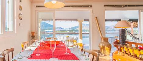 Comedor con una mesa dispuesta para dos, con vista a un cuerpo de agua y montañas al fondo.