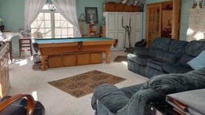 A living room with a pool table, a sofa, a ceiling fan, and a window with curtains. - Local Events House on Lake near HPU, High Point Markets, NC Hospitals  (High Point)