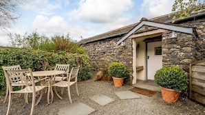 A stone building with a door, a small porch, and a table with chairs. There are potted plants and a gravel path. - Shepherds Cottage (Ambleside)