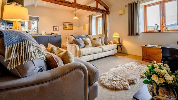 A cozy living room with a sofa, a coffee table, and a rug. There is a wooden beam structure on the ceiling, a window with curtains, and a lamp on a side table. - Shepherds Cottage (Ambleside)