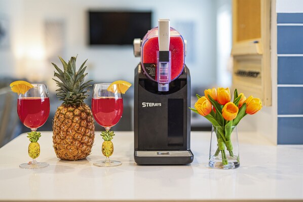 Una zona de bar con dispensador de bebidas, vasos con adornos de frutas y un jarrón con flores.
