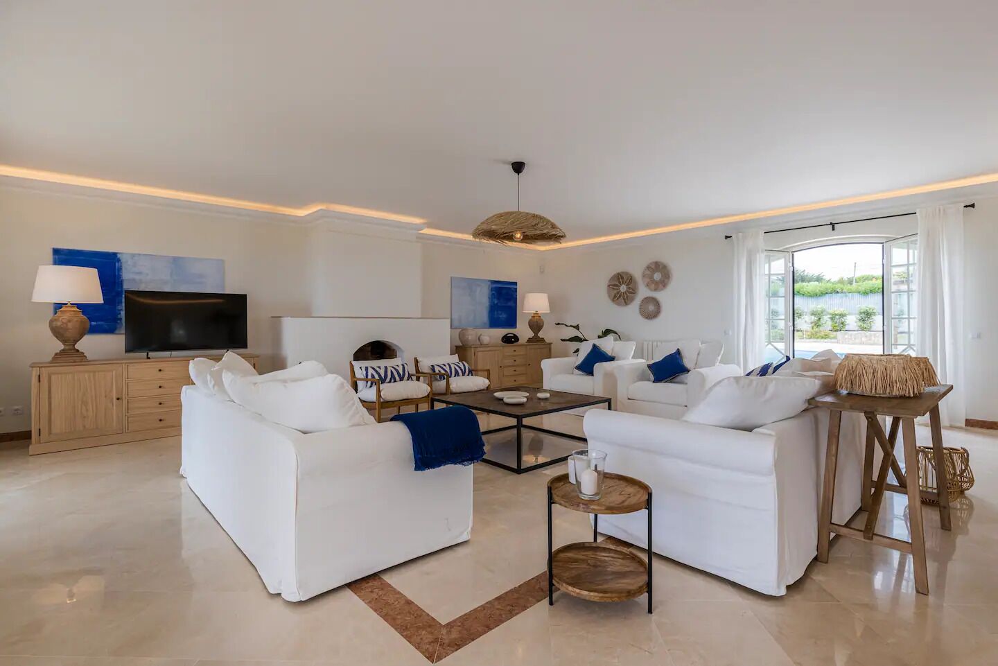 Ein modernes Wohnzimmer mit weißen Sofas, einem Couchtisch und einem Fernseher. Es gibt ein großes Fenster mit Vorhängen, eine Deckenleuchte und ein dekoratives Wandbild.