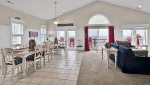 A spacious living room with a high ceiling, a dining area with white chairs and a table, and a large window with a view of the ocean. - Hot Tub, BeachFront, Private, Hammocks on the Beach (Surf City)
