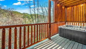 Una terraza de madera con un jacuzzi, barandillas de madera y vistas a árboles y colinas.