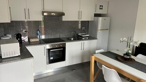 A modern kitchen with white cabinets, a stainless steel refrigerator, a built-in oven, and a dining area with a wooden table and white chairs. - Retiro Termal no Centro do Vulcão das Furnas (Furnas)