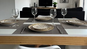 A modern dining area with a wooden table set for four, surrounded by white chairs. A black sofa and a television are visible in the background. - Retiro Termal no Centro do Vulcão das Furnas (Furnas)