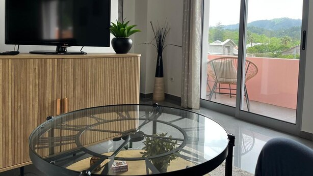 A modern living room with a flat-screen TV, a glass-top coffee table, a wooden cabinet, a potted plant, and a view of a balcony with outdoor furniture. - Retiro Termal no Centro do Vulcão das Furnas (Furnas)
