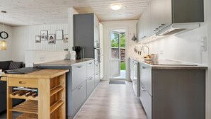 Eine moderne Küche mit grauen Schränken, einer Holzarbeitsplatte und einem eingebauten Backofen. Auf der Arbeitsplatte befinden sich eine Kaffeemaschine, ein Wasserkocher und ein Toaster. Die Küche hat Holzboden und einen Blick auf den Garten durch eine offene Tür.