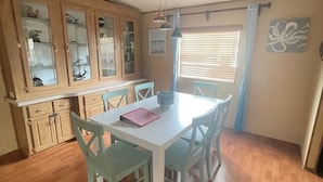 Un coin repas avec une table blanche et des chaises bleues, des meubles en bois dotés de portes en verre et une fenêtre avec des stores.