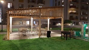 Eine hölzerne Gartenpergola mit Tischen und Stühlen, umgeben von einer Rasenfläche und einem Gebäude im Hintergrund.