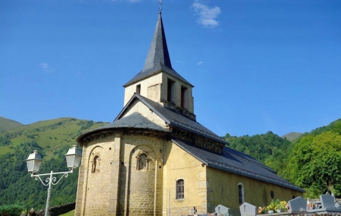 Església amb un campanar alt i apuntat i una torre de campanes arrodonida, envoltada per turons verds i exuberants.