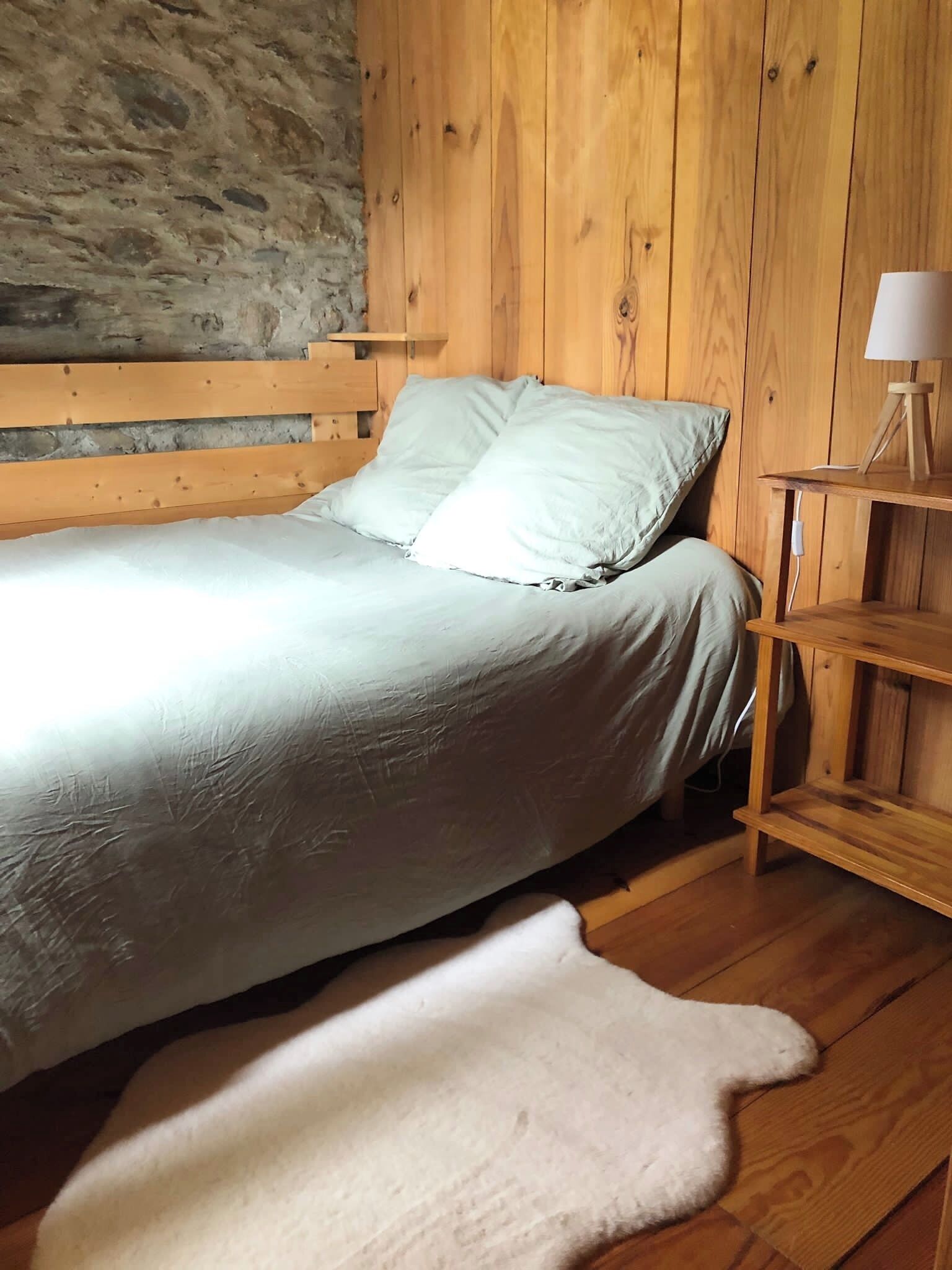 Una cama de madera con un colchón y almohadas blancos, una alfombra blanca y una mesita de noche con una lámpara.