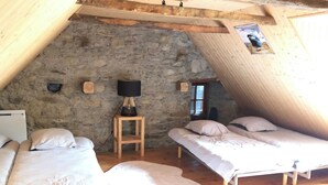 A cozy attic bedroom with two beds, a small table with a lamp, and a stone wall.