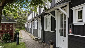 Une rangée de maisons en bois sombre, ornées de boiseries blanches, portant les numéros 34, s’étend le long d’un chemin pavé, bordé d’une clôture en bois et de verdure.
