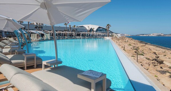 A poolside area with lounge chairs and umbrellas, a clear view of the sea, and a rocky shoreline. - Convenient Central 3BR Apt Near Bus Terminus (St. Paul's Bay)