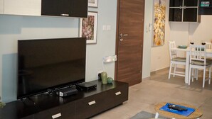 A modern living room with a flat-screen TV, a dark entertainment unit, a dining area with a table and chairs, and a kitchen area with cabinets and a microwave. - Convenient Central 3BR Apt Near Bus Terminus (St. Paul's Bay)