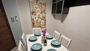 A dining area with a white table set for four, teal plates, white chairs, a vase with purple flowers, and a painting of flowers on the wall. - Convenient Central 3BR Apt Near Bus Terminus (St. Paul's Bay)