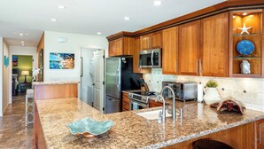 A modern kitchen with granite countertops, stainless steel appliances, wooden cabinets, and a large island with bar stools. - Fully Loaded 1 Bedroom 2 Bathroom Condo in Kihei, Maui!  (Kihei)