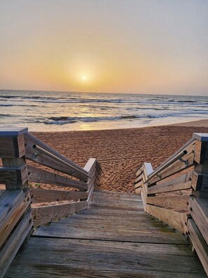 A wooden boardwalk leading to a beach with the sun setting in the background.