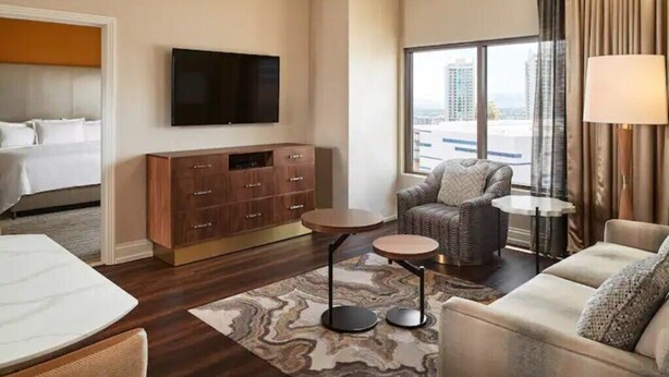 A hotel room with a bed, a television, a seating area with a sofa and armchairs, a small table, and a large window with a view of buildings.