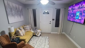 A living room with a yellow sofa, a brown wicker chair, a white furry rug, a ceiling fan, and a TV on the wall. - Cozy Home on RGV (Alton)
