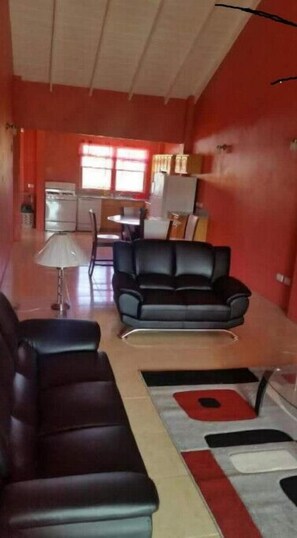 Un salon avec un canapé en cuir noir, une table à manger avec des chaises, et un coin cuisine à l’arrière-plan.