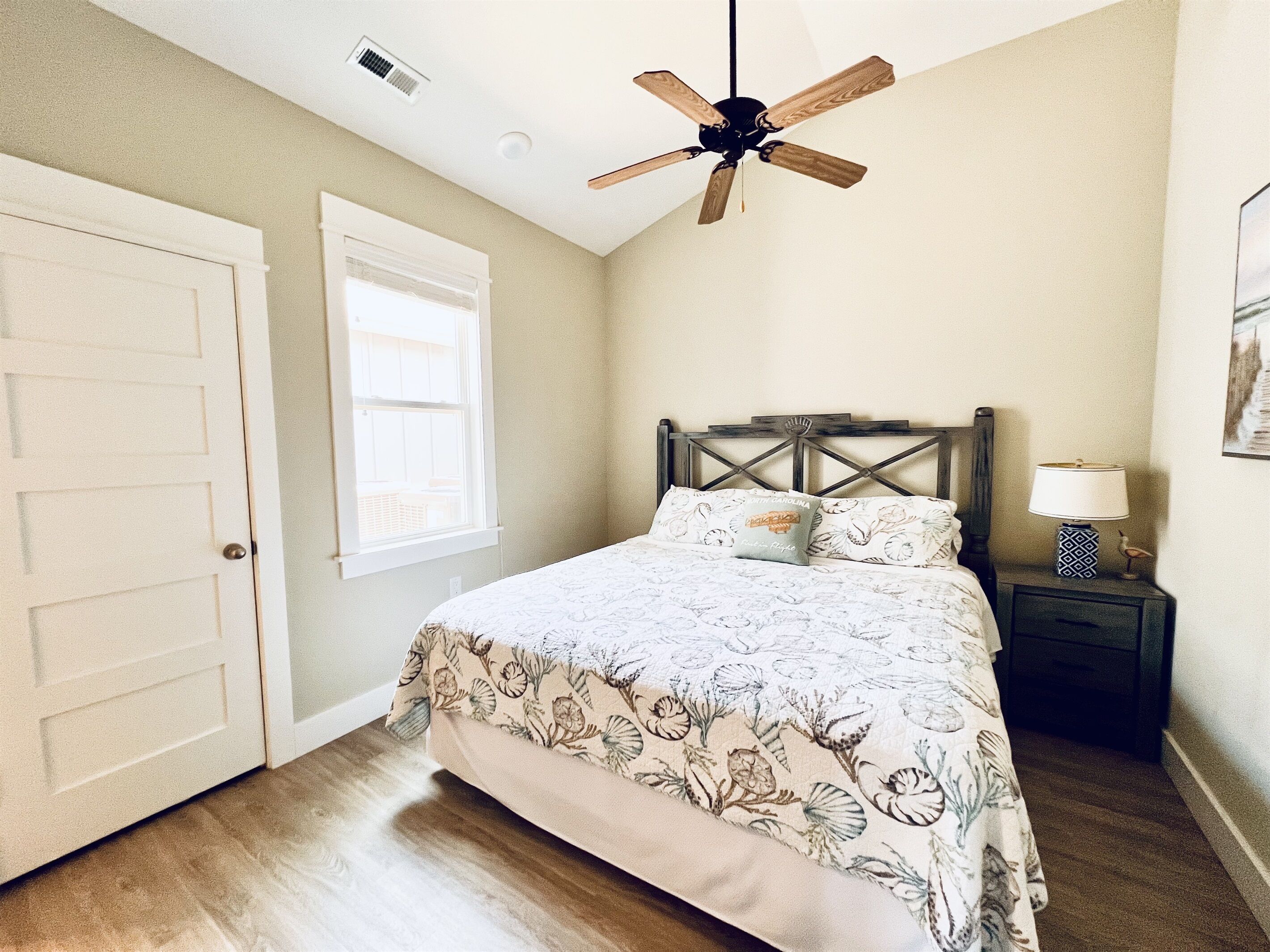 A bedroom with a bed, a ceiling fan, a nightstand with a lamp, and a window.