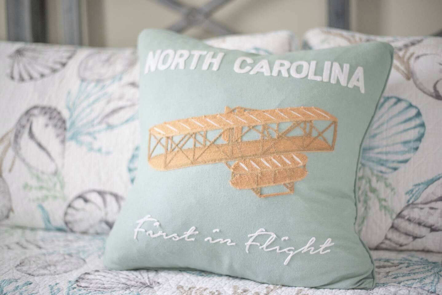 A pillow with embroidered text and a biplane design, placed on a patterned fabric.