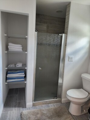 A bathroom with a glass shower door, a toilet, and shelves with folded towels.